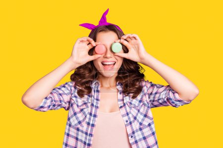 Portrait of nice cheerful girlish crazy attractive funny wavy-haired lady having fun holding in hands two macarons closing covering eyes isolated over violet background.