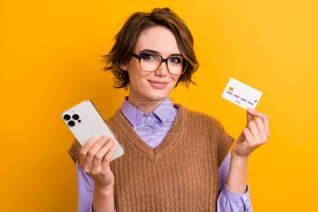 Photo of smart attractive girl dressed knitwear waistcoat in glasses hold smartphone credit card isolated on yellow color background.