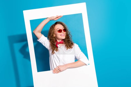 Foto-Porträt von netten jungen Dame suchen leeren Raum Sonnenbrille sofortige Fotorahmen tragen trendige weiße Kleidung isoliert auf blauem Hintergrund.