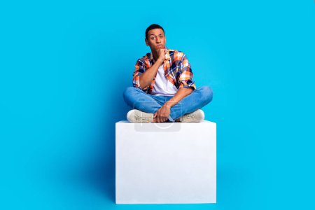Full size photo of nice young man sit podium think empty space wear shirt isolated on blue color background.