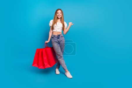 Retrato de longitud completa de hermosa chica mantenga bolsas dedo directo espacio vacío desgaste superior aislado sobre fondo de color azul.