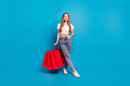 Retrato de longitud completa de hermosa chica mantenga bolsas de centro comercial espacio vacío desgaste superior aislado sobre fondo de color azul.