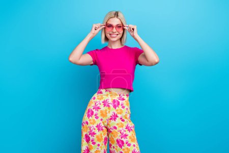 Foto retrato de encantadora joven toque gafas de sol vacaciones de verano vestido con estilo ropa rosa aislado sobre fondo de color azul.
