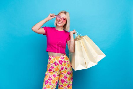 Foto retrato de atractiva mujer joven gafas de sol mantenga bolsas de compras vestidas con estilo ropa rosa aislado en el fondo de color azul.