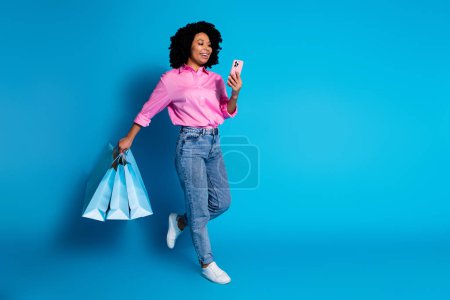 Full body portrait of pretty young woman use smart phone hold bags empty space wear pink shirt isolated on blue color background.