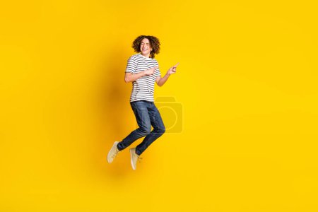 Full size portrait of nice young man jump point fingers empty space wear striped t-shirt isolated on yellow color background.