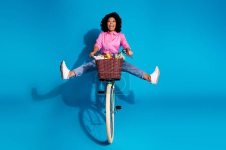 Full size photo of eccentric cereless woman wear pink shirt driving bicycle with flowers in bucket isolated on blue color background.