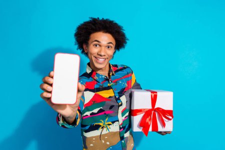 Photo portrait of handsome young guy hold gadget show screen present box dressed stylish print garment isolated on blue color background.