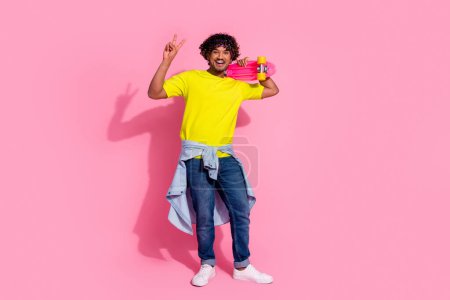 Photo de drôle homme joyeux porter élégant vêtements jaunes tenir skateboard v-signe isolé sur fond de couleur rose.