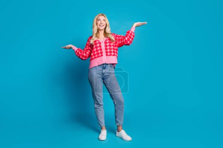 Full size photo of pleasant person dressed knitwear jumper jeans look at best variant on arm empty space isolated on blue color background.