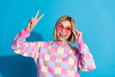 Photo portrait of pretty young girl touch sunglass celebrate party show v-sign wear trendy pink outfit isolated on blue color background.