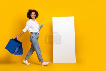 Ganzkörpergröße Ansicht der schönen trendigen fröhlichen Mädchen mit Gadget tragen Güter isoliert über helle gelbe Farbe Hintergrund.