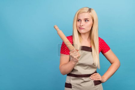 Photo portrait housewife wearing apron keeping rolling pin looking copyspace unsure isolated pastel blue color background.