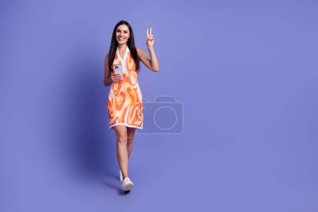 Full lengh photo of pretty lady wear dress showing v-sign communicating gadget emtpy space isolated purple color background.