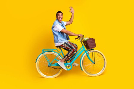 Full length photo of friendly guy dressed jeans waistcoat driving cycle to empty space say hi isolated on vibrant yellow color background.