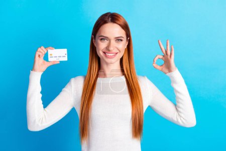 Foto-Porträt von attraktiven jungen Frau halten Kreditkarte okey gekleidet stilvolle weiße Kleidung isoliert auf blauem Hintergrund.