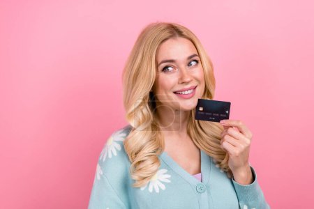 Porträt der netten Person mit lockigem Haar tragen blaue Strickjacke halten Kreditkarte Blick auf Promo leeren Raum isoliert auf rosa Hintergrund.