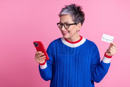 Photo de charmante femme âgée gaie porter des vêtements bleus tendance tenir virement de transaction par carte bancaire isolé sur fond de couleur rose.