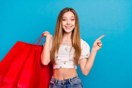 Foto de positiva chica feliz mantenga bolsas de compras demuestran punto vacío espacio aislado color azul fondo.