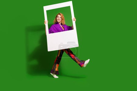 Photo portrait of lovely young lady walk hold instant photo frame dressed stylish violet clothes isolated on green color background.