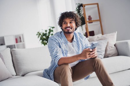 Foto von gut aussehenden fröhlichen Kerl kariertes Hemd genießen Wochenende SMS-Schreiben modernes Gerät drinnen Zimmer zu Hause Haus.