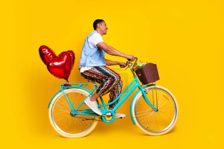 Full size photo of cheerful nice man dressed denim vest ride cycle with balloons look empty space isolated on yellow color background.