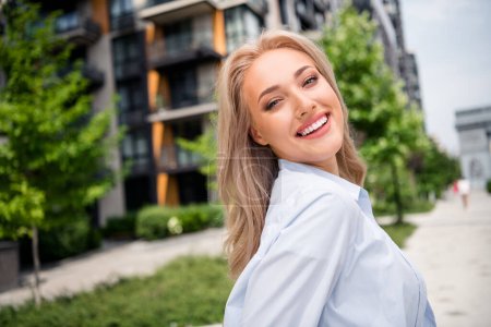 Portrait of attractive dreamy young adorable strolling girl wearing blue shirt posing near modern design residential complex outdoors.