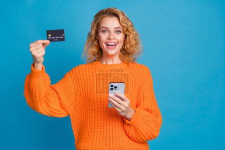 Photo positive jolie femme porter des vêtements orange élégant tenir la transaction de transfert de carte de crédit bancaire isolé sur fond de couleur bleue.