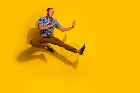 Full length photo of handsome good mood guy wear print shirt jumping high kicking empty space isolated yellow color background.