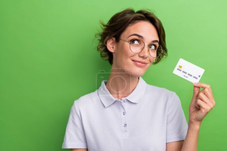 Photo of lovely girl wear trendy clothes hand hold bank credit card look up empty space offer banner isolated on green color background.