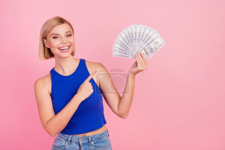 Portrait of nice young girl direct finger dollar bills wear blue top isolated on pink color background.
