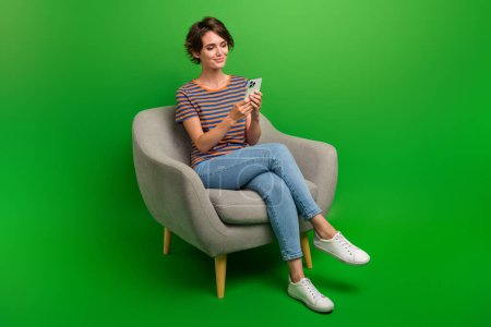 Retrato de tamaño completo de encantadora hermosa chica sentarse silla acogedora uso de mensajes de texto de teléfonos inteligentes aislados en el fondo de color verde.