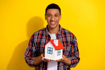 Portrait de bonne humeur satisfait gars avec une coiffure courte porter chemise à carreaux tenant petite maison dans les mains isolées sur fond de couleur vive.