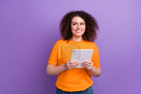 Photo of funky dreamy woman wear orange t-shirt chatting apple samsung modern device looking empty space isolated purple color background.