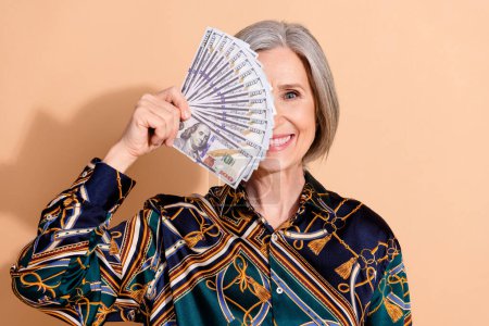 Photo of satisfied funky retired person with bob hairdo wear print blouse dollars cover eye smiling isolated on beige color background.
