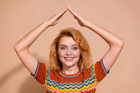 Portrait of good mood funky girl with ginger hairdo wear knit t-shirt palms show roof over head isolated on beige color background.