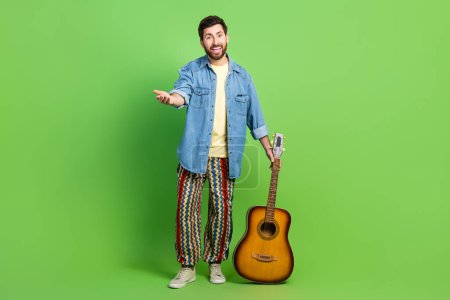 Ganzes Foto von fröhlichen positiven Kerl gekleidet Jeans-Shirt lädt Sie ein, Gitarrenmusik genießen isoliert grüne Farbe Hintergrund.