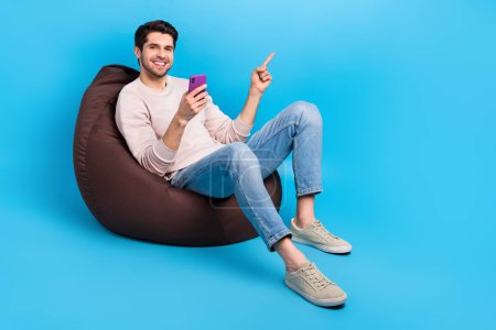 Full length photo of nice guy wear pullover sit on pouf indicating at eshop empty space hold smartphone isolated on blue color background.