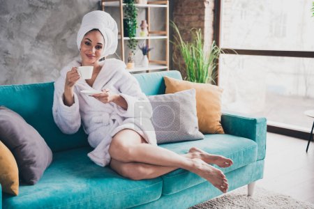 Portrait photo de jolie jeune femme assise canapé tenir tasse de café tête enveloppée serviette habillée peignoir de bain intérieur profiter week-end.