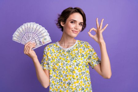 Photo of pretty young woman hold money show okey symbol wear t-shirt isolated on violet color background.