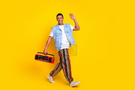 Full length photo of cheerful guy dressed jeans waistcoat hold boombox say hi walk empty space isolated on yellow color background.