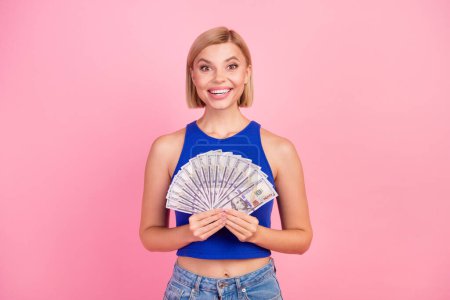 Foto de mujer bastante joven sostiene billetes de dólar usan top azul aislado sobre fondo de color rosa.
