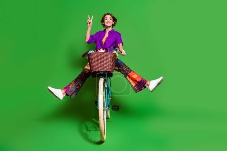 Photo portrait of funny young woman wear purple shirt and brown bob haircut riding bicycle showing v sign isolated on green color background.