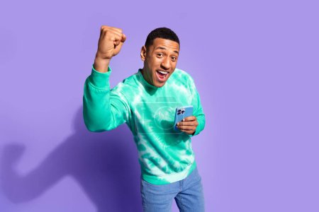 Photo of attractive lucky guy wear teal sweatshirt winning game modern gadget emtpy space isolated purple color background.