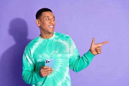 Photo of attractive funny guy wear teal sweatshirt typing modern gadget showing emtpy space isolated purple color background.
