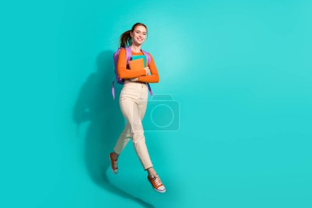 pleine taille photo de fille intelligente habillé orange chemise sac à dos tenir livre volant à l'université vide espace isolé sur fond de couleur sarcelle.