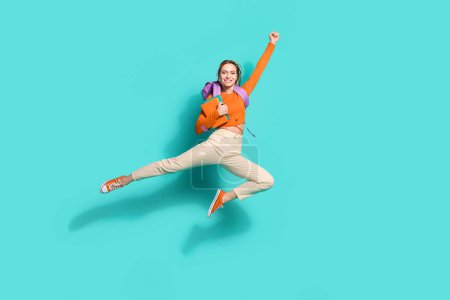 Photo pleine grandeur de fille optimiste habillée sac à dos chemise orange sur l'épaule livre volant levant poing vers le haut isolé sur fond de couleur sarcelle.