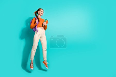 Photo pleine grandeur de fille optimiste habillé sac à dos chemise orange sur l'épaule livre regarder espace vide isolé sur fond de couleur sarcelle.