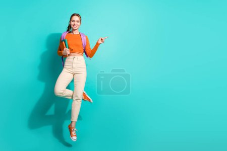 pleine taille photo de fille intelligente habillé orange chemise sac à dos tenir livre volant indiquant espace vide isolé sur fond de couleur sarcelle.