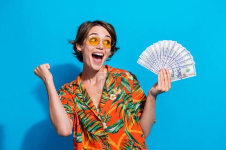 Photo de dame riche réjouie lever poing tenir billets de dollar regarder vide annonce de l'espace isolé sur fond de couleur bleue.